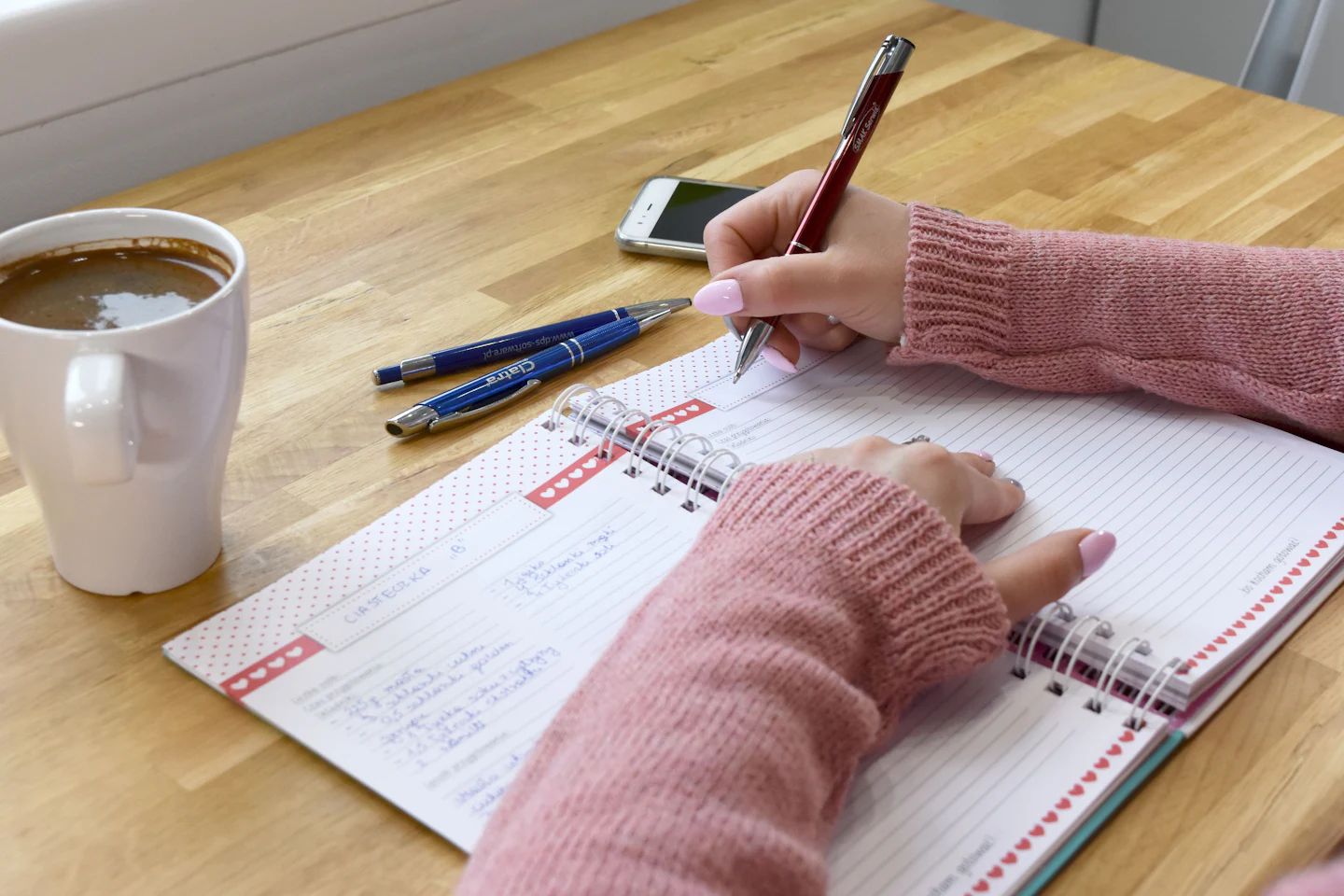 Hands writing in a spiral planner beside a cup of coffee on a wooden table