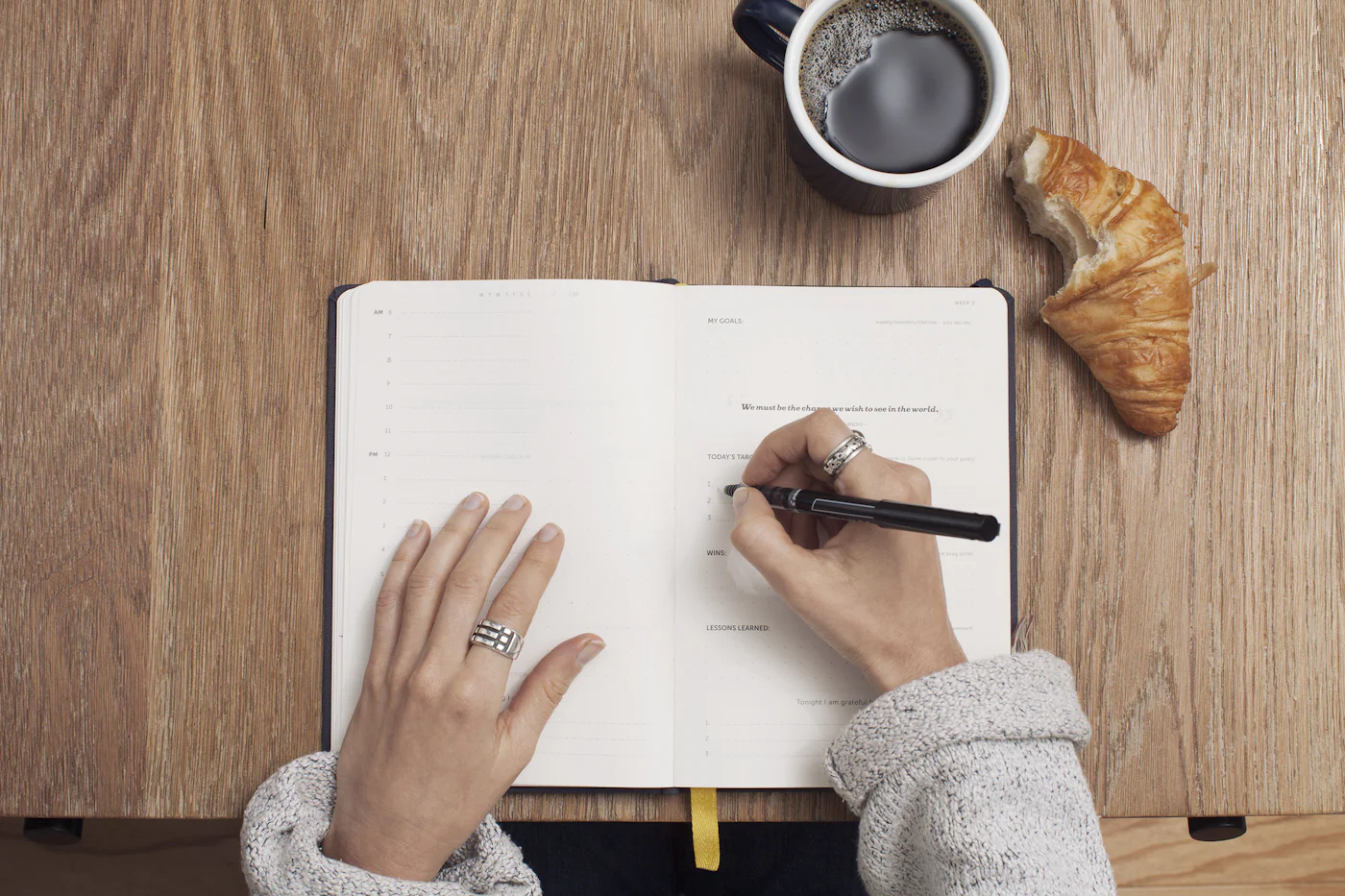 A person writing in an open notebook beside a coffee and a croissant