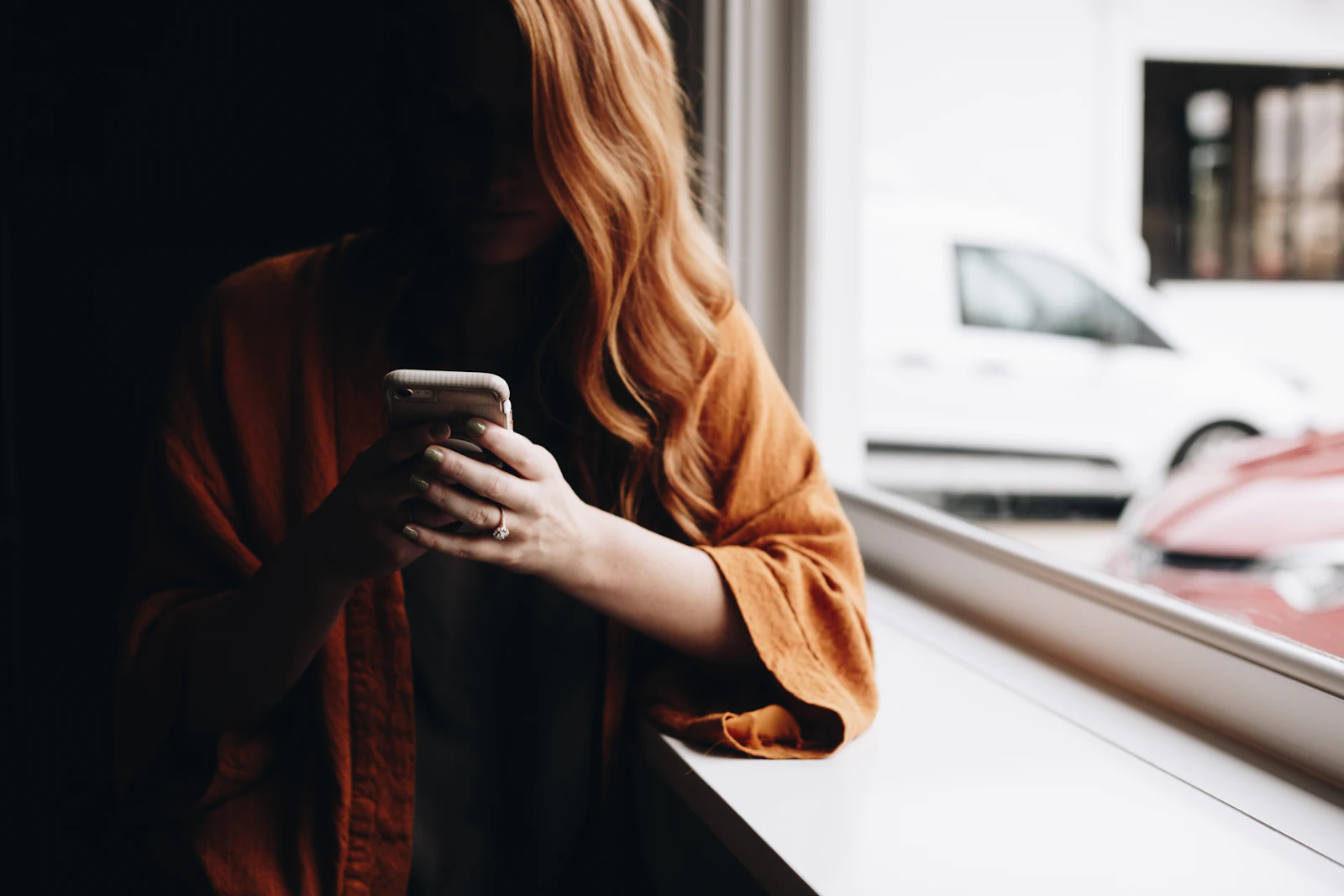 A woman in an orange cardigan texting at a window, her face out of frame
