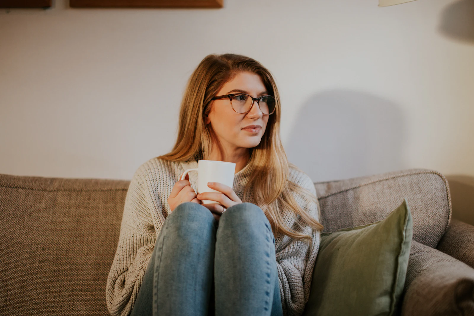 A woman in glasses sitting on a sofa with a mug, looking thoughtfully into the distance