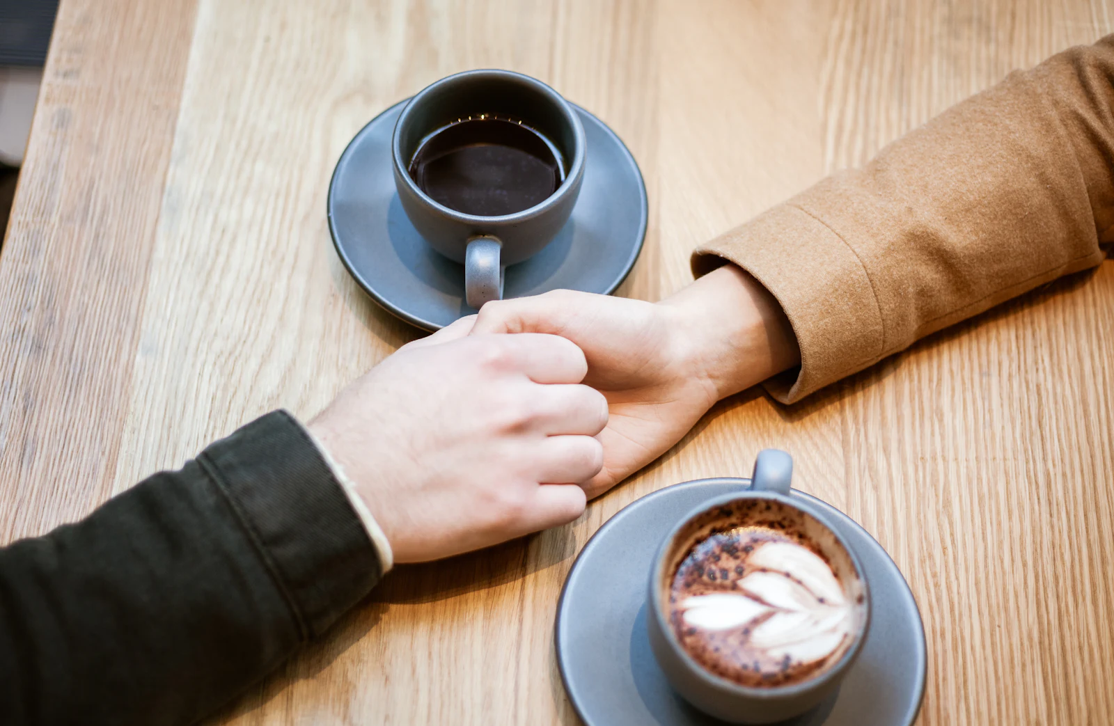 Two people holding hands on a wooden table between two cups of coffee