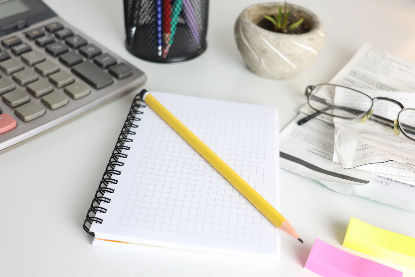 A desk with a calculator, a spiral notebook with a pencil, glasses and sticky notes