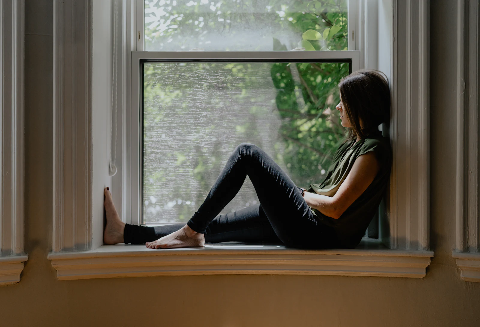 A woman sitting on a window sill, looking out at the trees