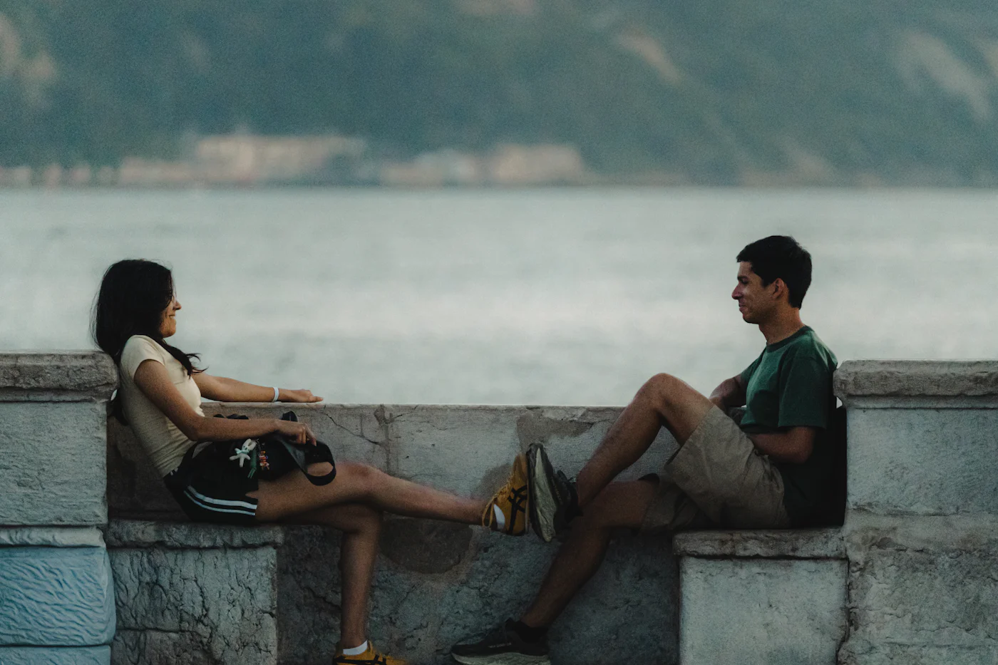A woman and a man sitting a little apart on a stone wall by the water