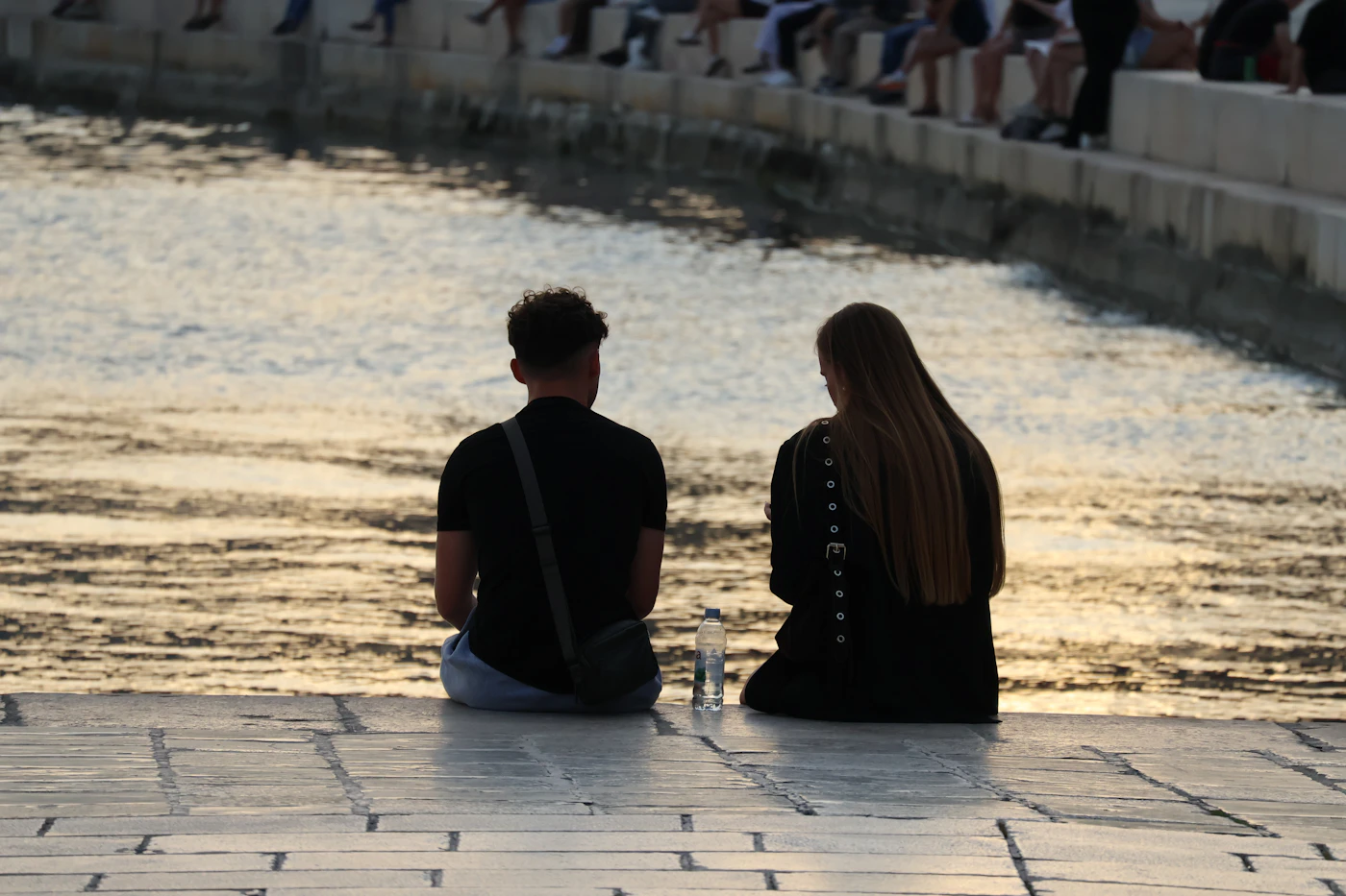 Two people sitting side by side on a stone step by sunlit water, seen from behind