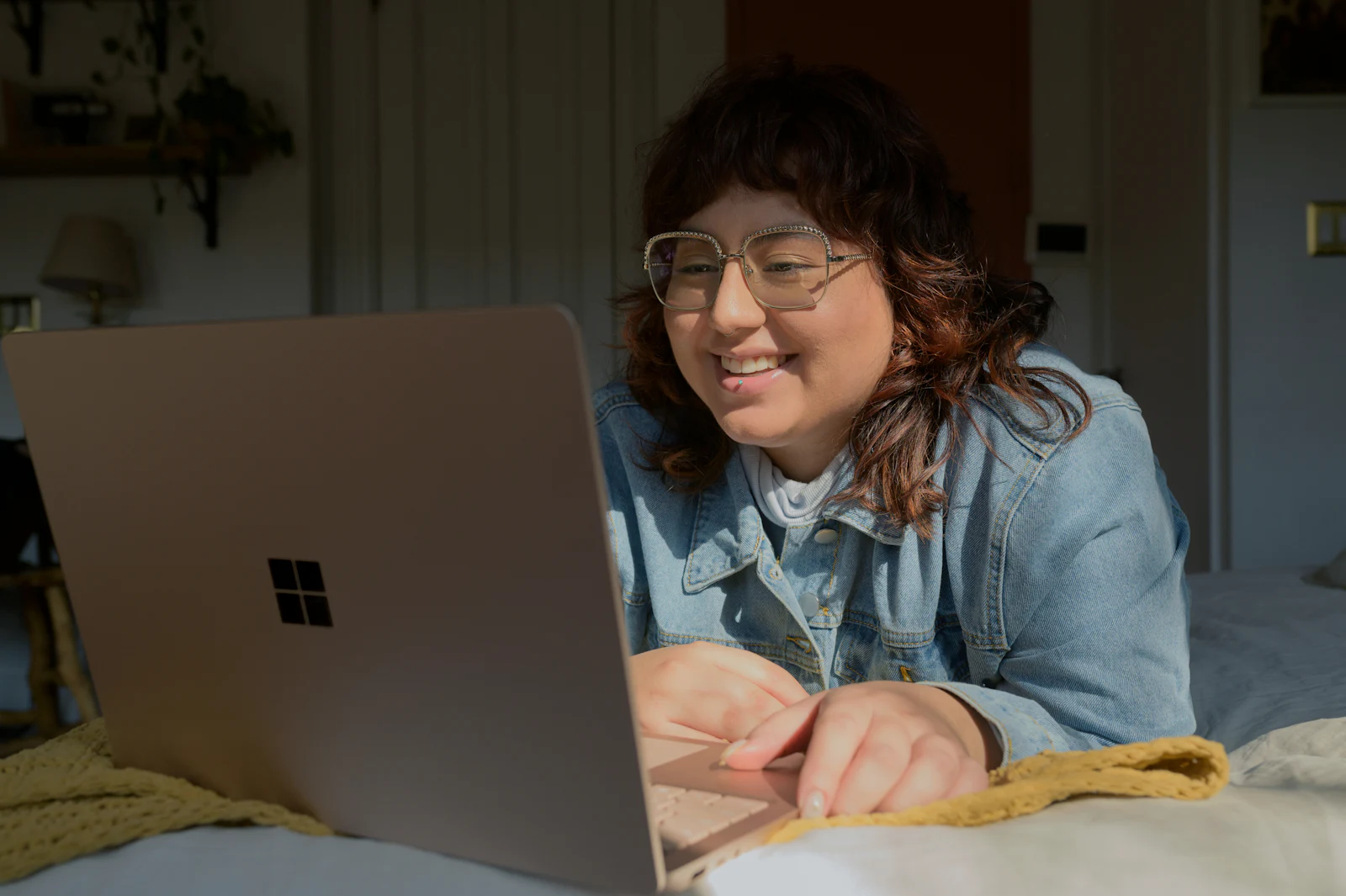 A woman in a denim jacket smiling at her laptop on a bed, mid conversation